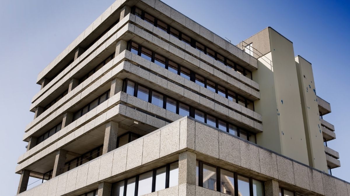 Brutalist-style concrete building with multiple stacked rectangular levels and large dark-framed windows, shown from a low angle against a clear blue sky
