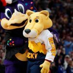 Two NBA mascots, including Rocky the Mountain Lion, hug on the court during a game with fans cheering in the background