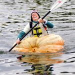 Person kayaking in a giant hollowed pumpkin on a lake during a pumpkin regatta.
