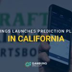 Person holding a smartphone displaying a sports prediction platform in front of a DraftKings Sportsbook sign.